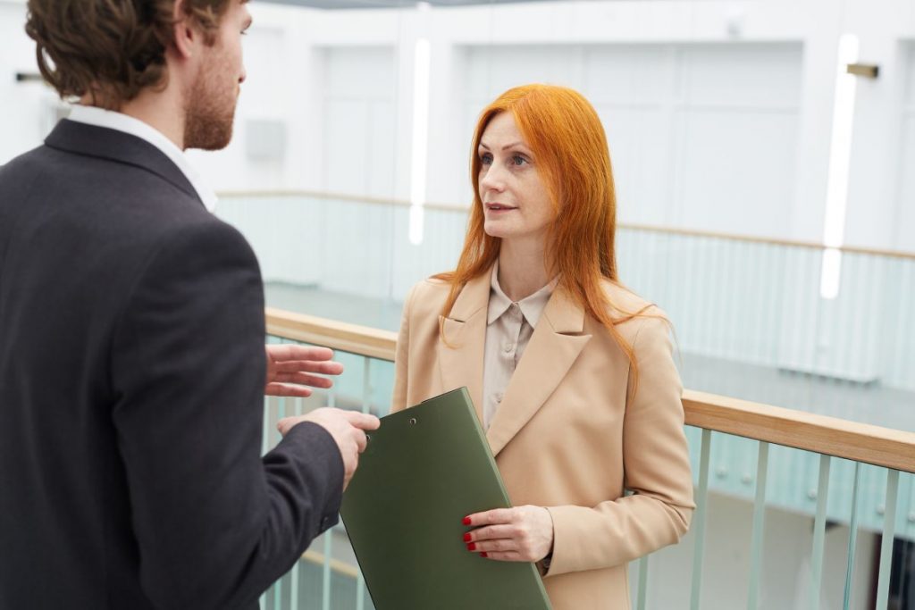 Waist up portrait of of red-haired female manager talking to colleague while standing on balcony in white office interior, copy space